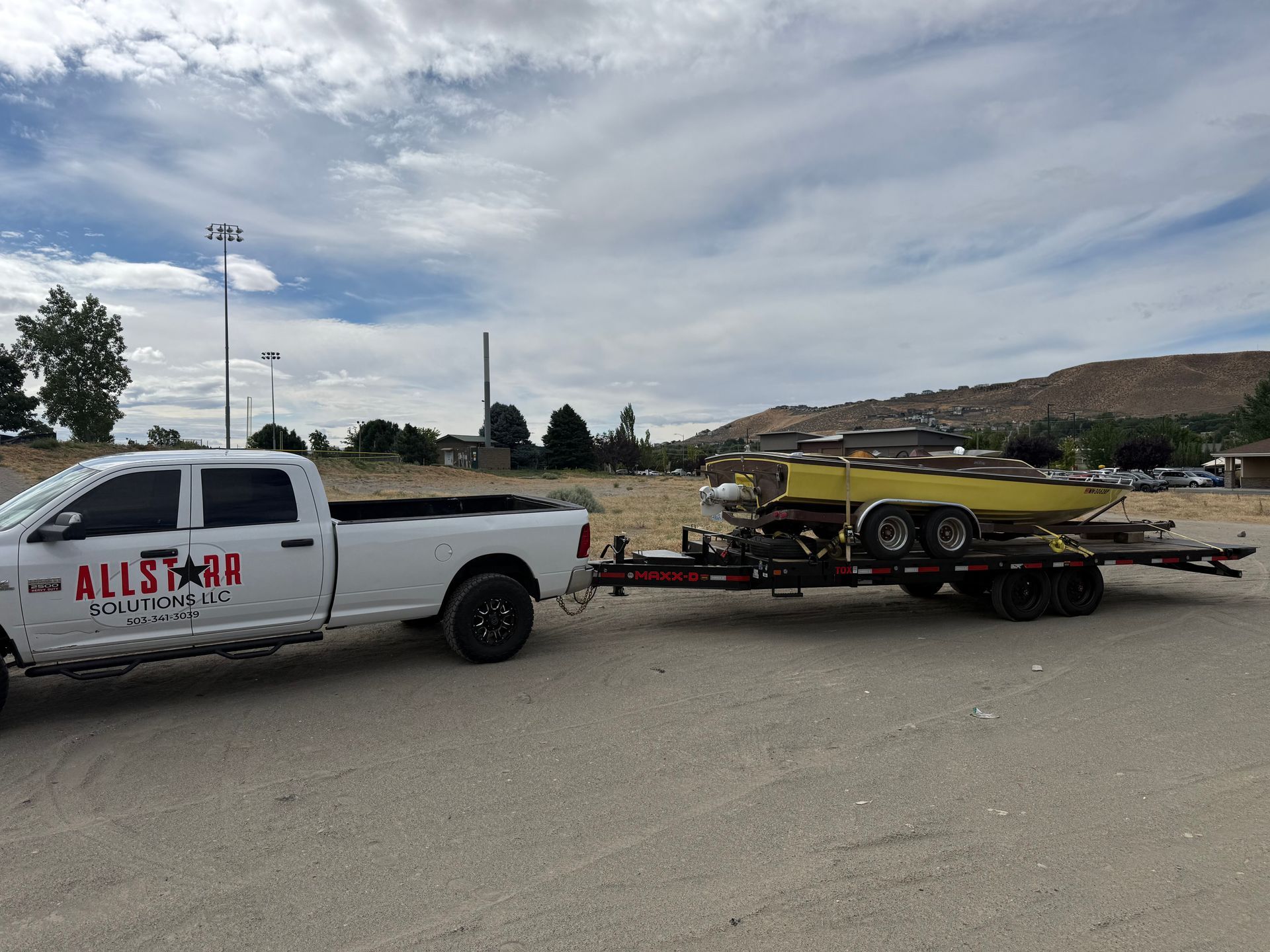 White pickup truck towing a flatbed trailer with yellow equipment on a roadside under a cloudy sky