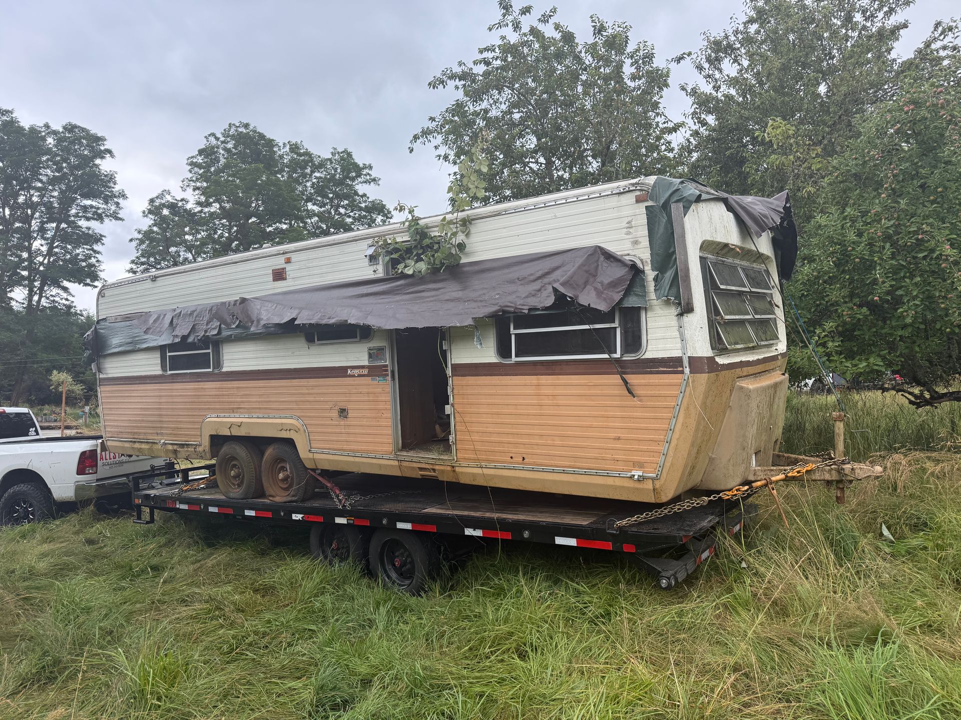 Damaged beige trailer home on a flatbed in a grassy field, with a torn roof and trees behind it