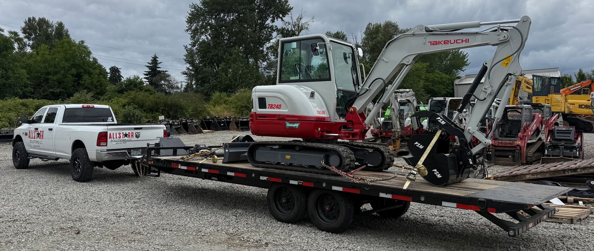 White pickup towing a flatbed trailer with compact excavators in a gravel equipment yard