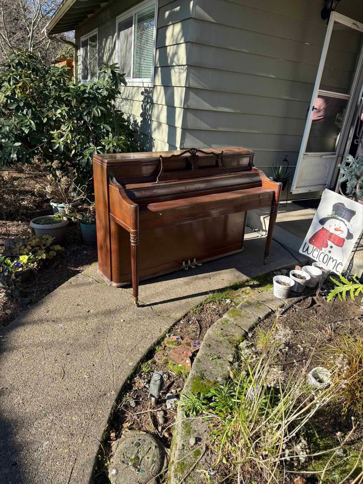 A wooden upright piano sits on a concrete walkway outside a house next to a snowman-themed garden sign.