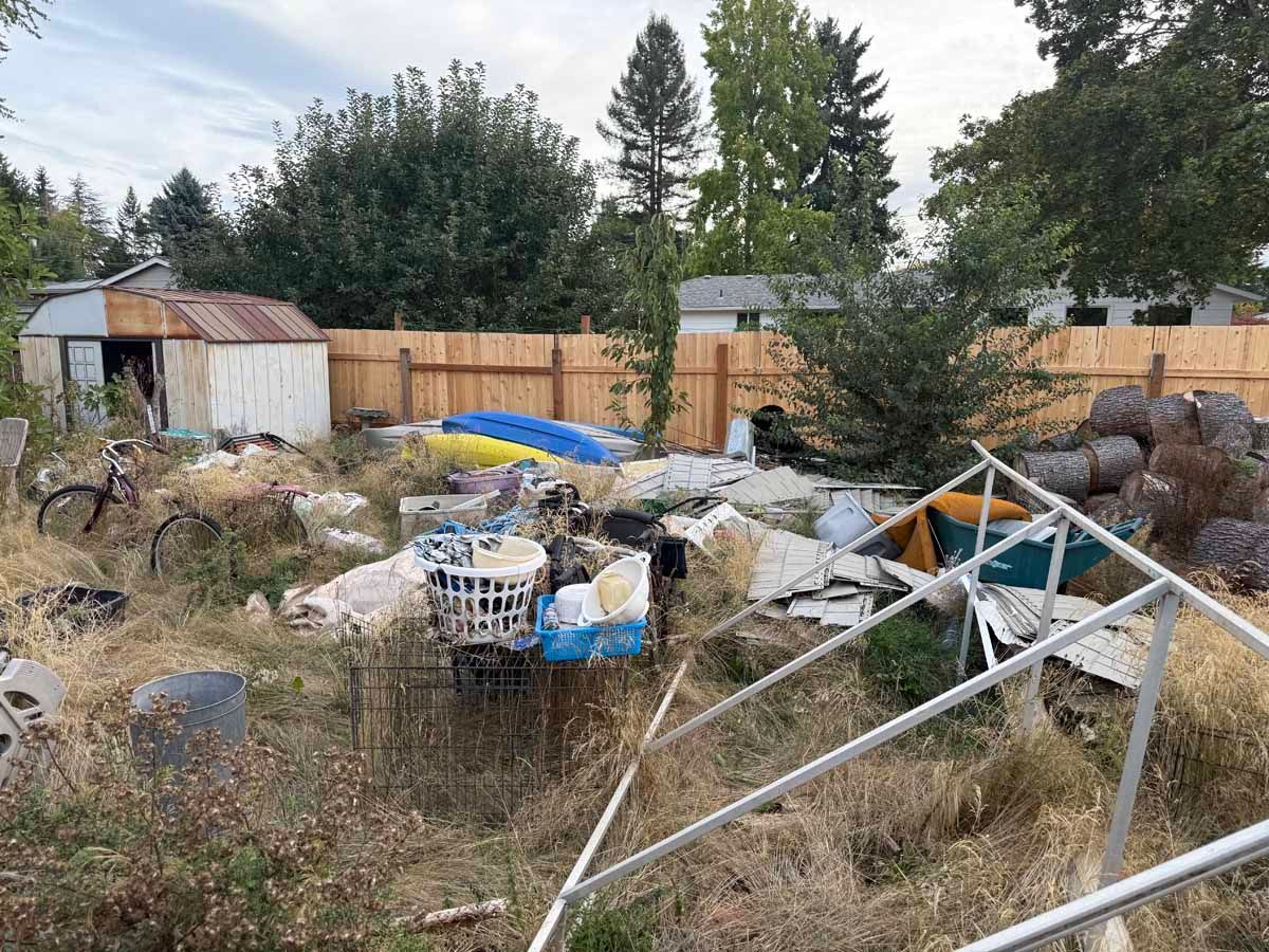 A messy backyard filled with scattered debris, a shed, a bicycle, and a metal frame structure against a wooden fence.
