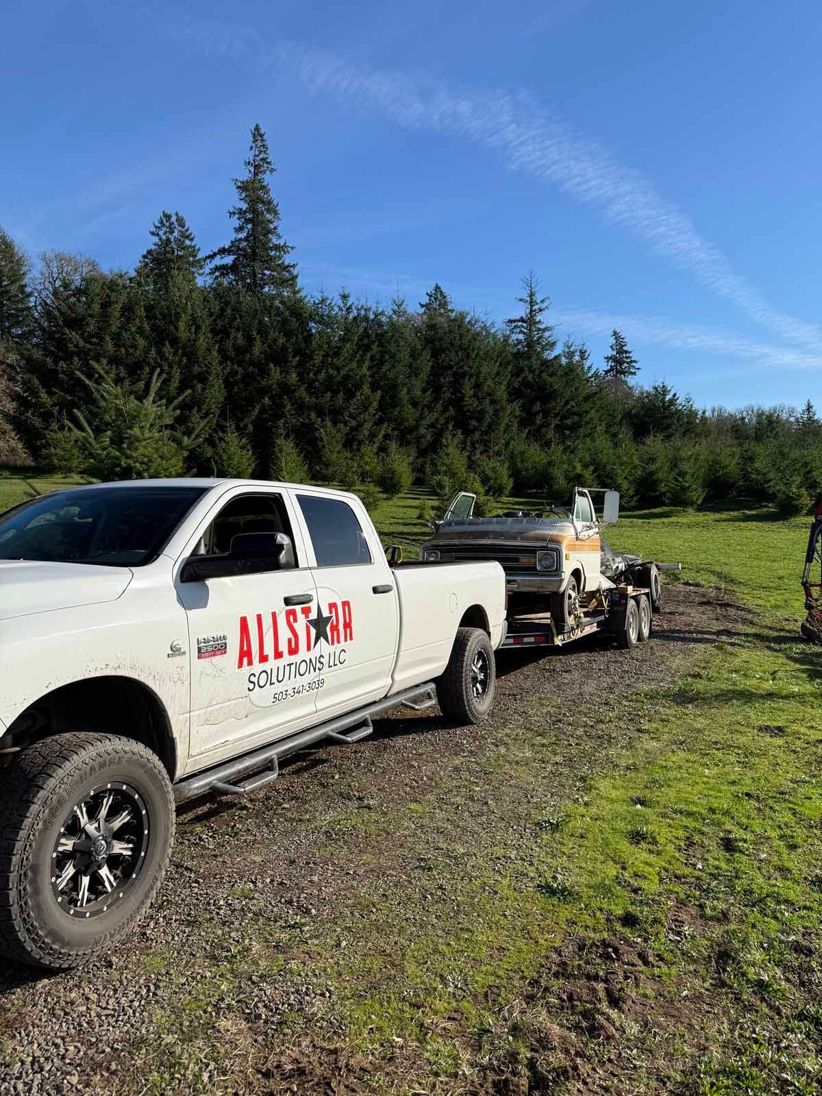 A white Allstar towing truck parked on a gravel path, hitched to a trailer carrying a small boat in a grassy, tree-lined field.