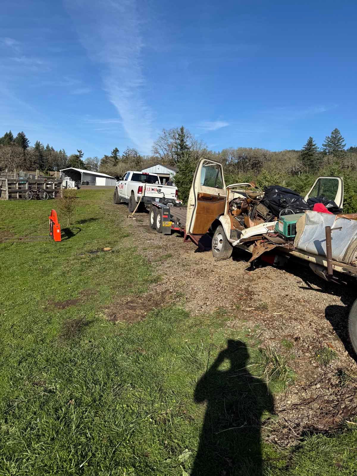 A white pickup truck tows a flatbed trailer loaded with a stripped, rusty vintage truck cab across a grassy field.