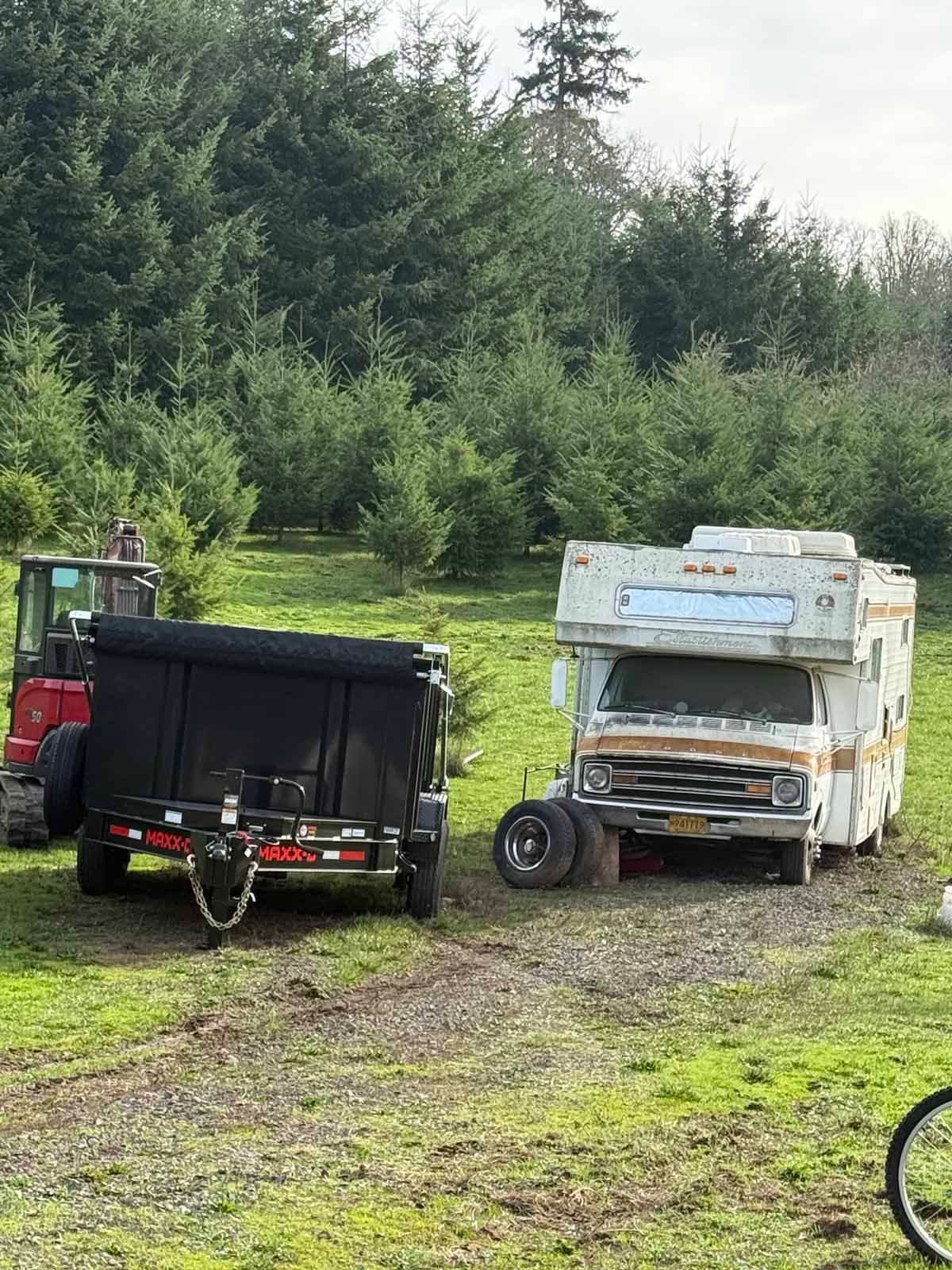 A red piece of heavy equipment pulling a black trailer next to a rusted white and orange camper in a field near a forest.