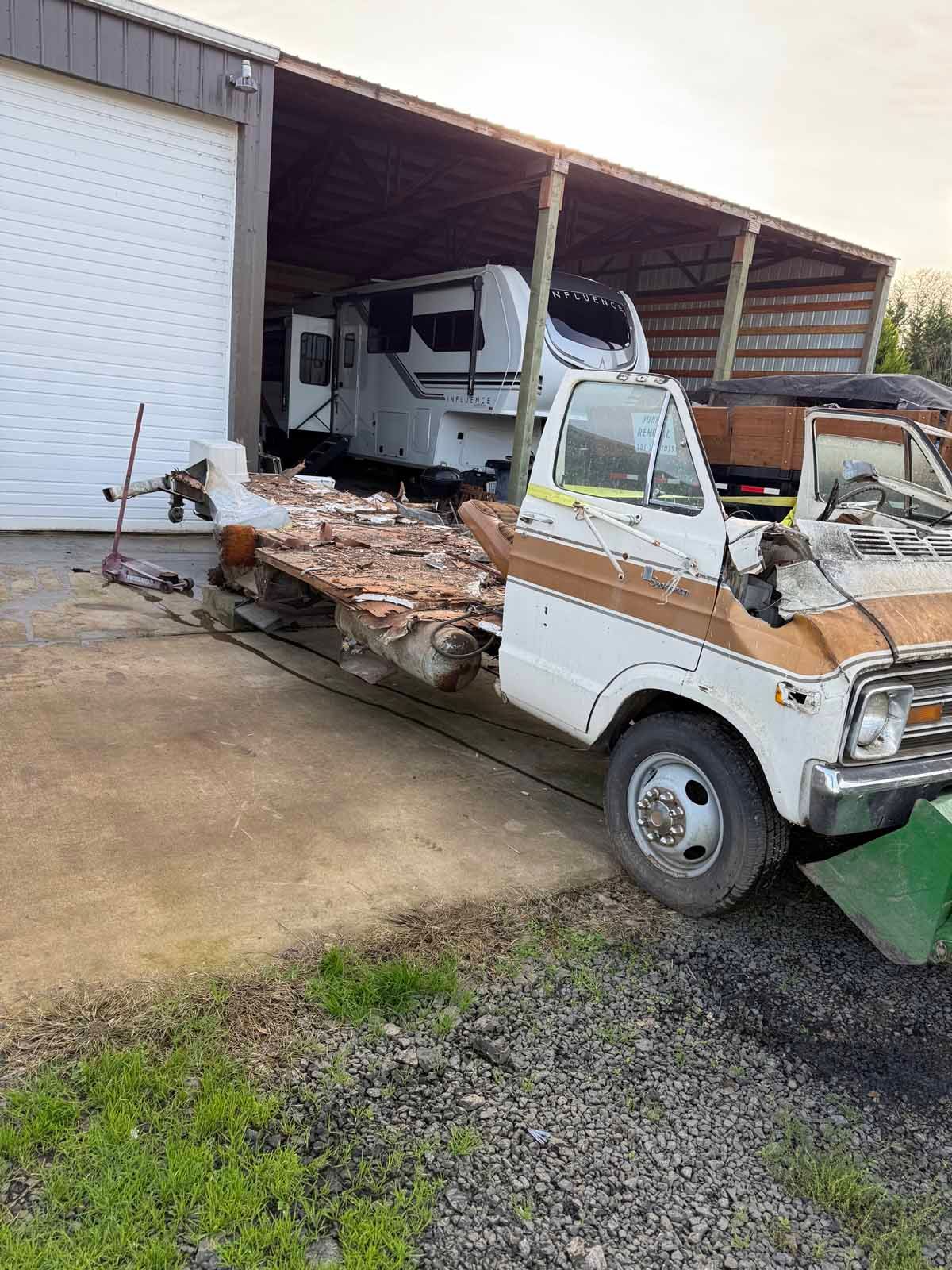 A white and tan cab-over truck frame parked outside a large metal barn with a camper in the background.