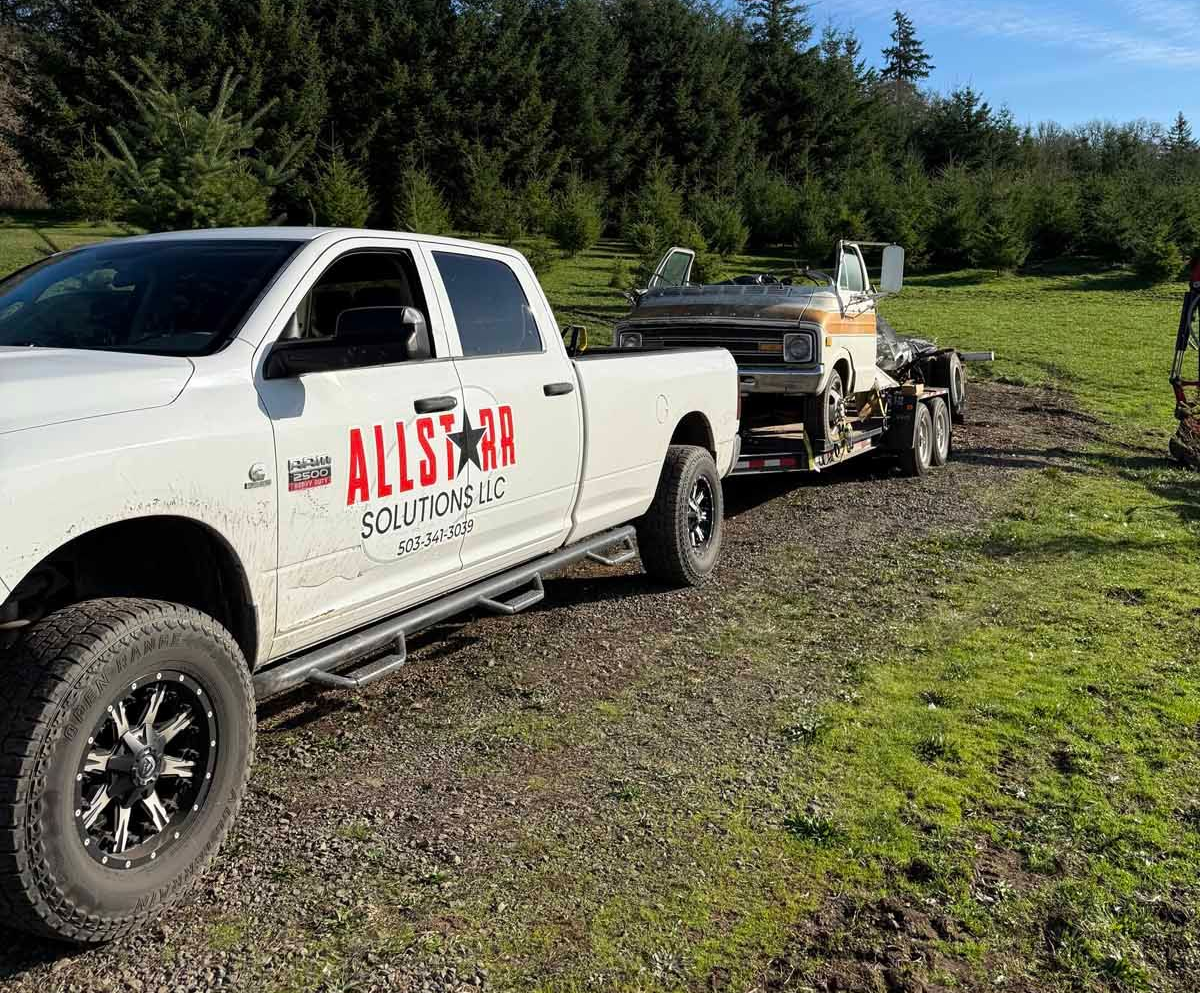 A white Allstar towing truck parked in a grassy field, pulling a trailer with a vintage vehicle on it.