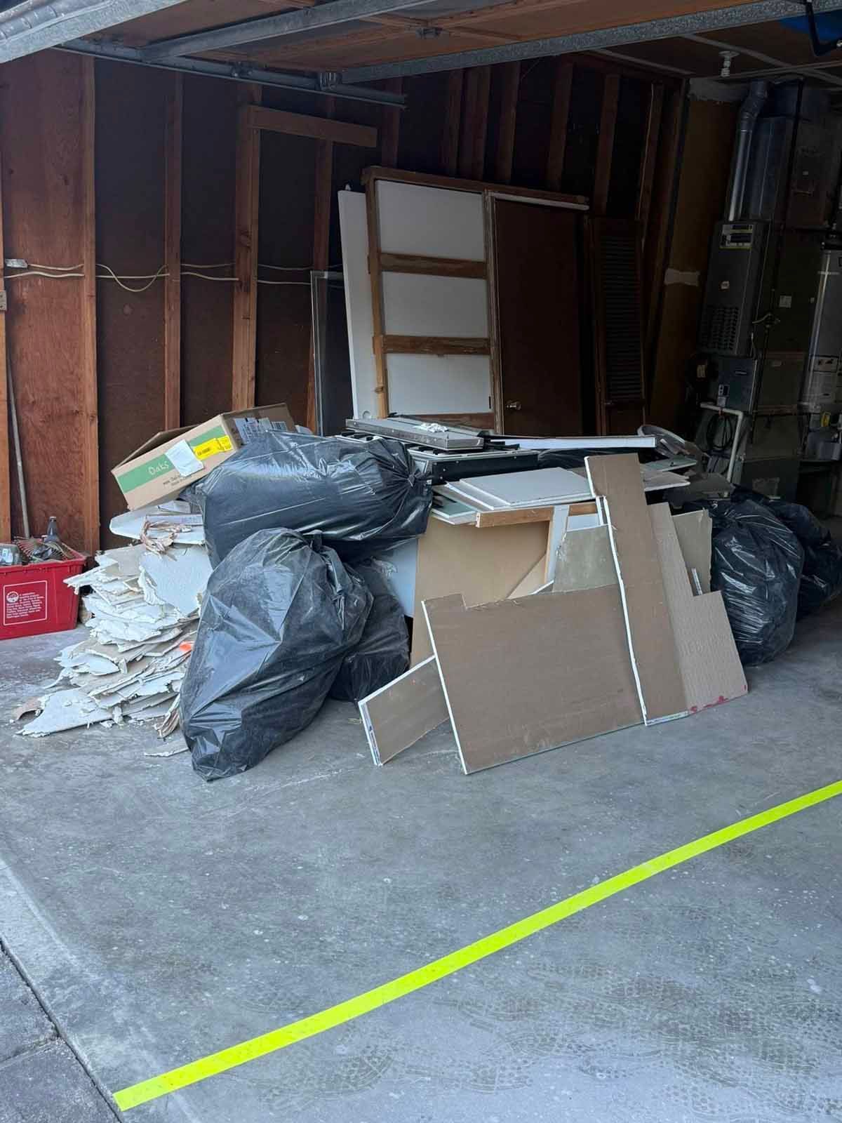 A garage corner filled with construction debris, including drywall scraps, black trash bags, and wood framing materials.