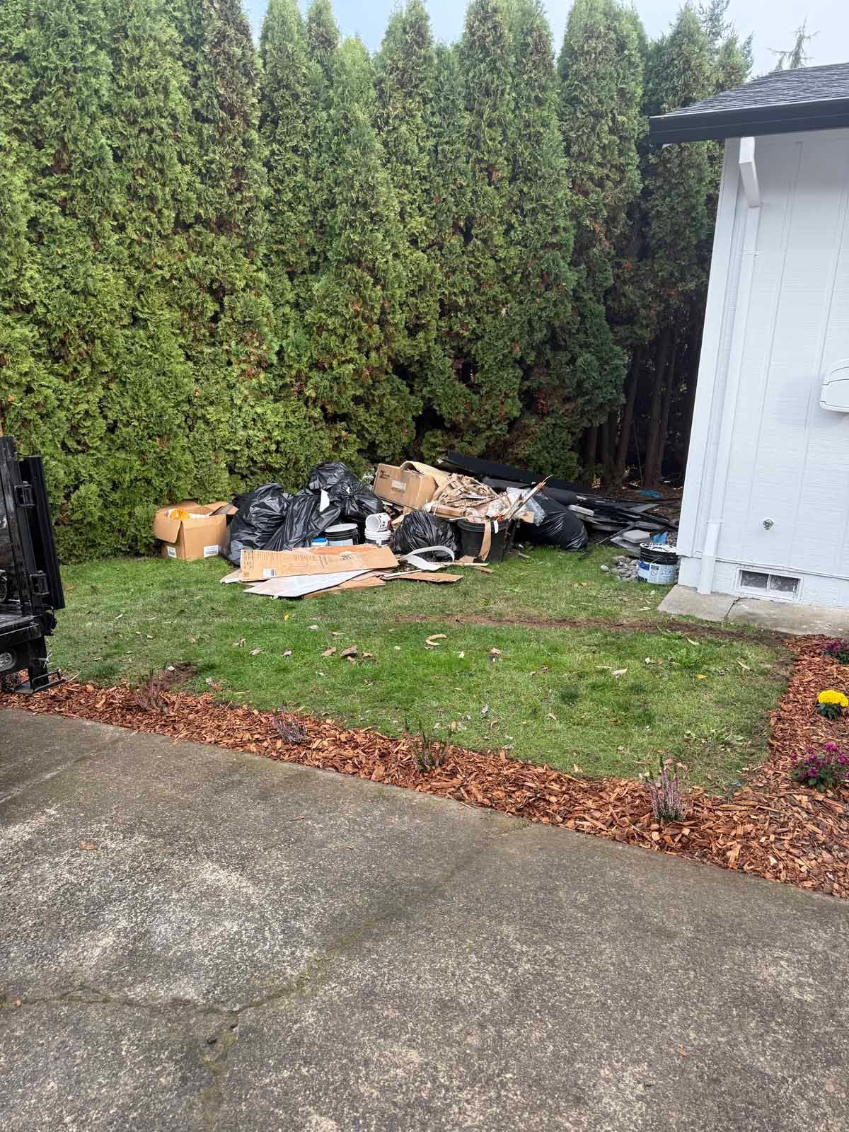 A pile of cardboard boxes and black trash bags sits on a lawn in front of tall evergreen trees next to a white house.