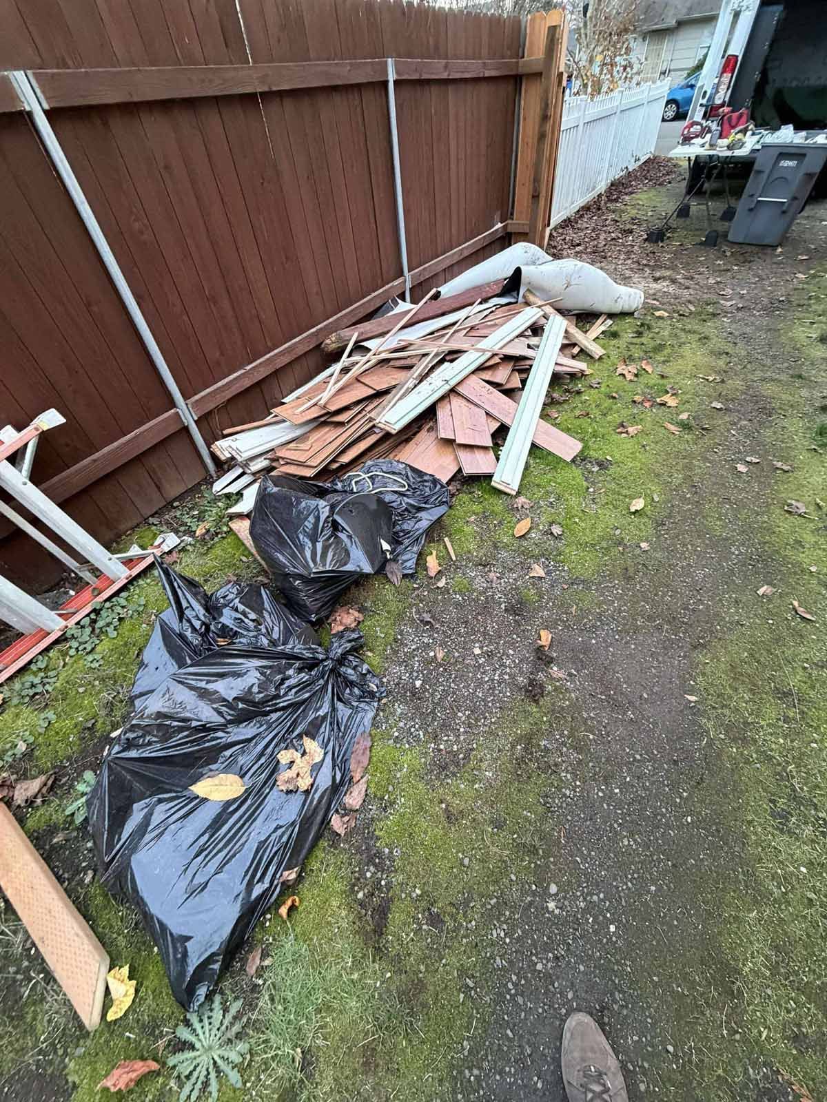 A pile of construction debris, including wood scraps and black trash bags, sits on a mossy yard next to a wooden fence.