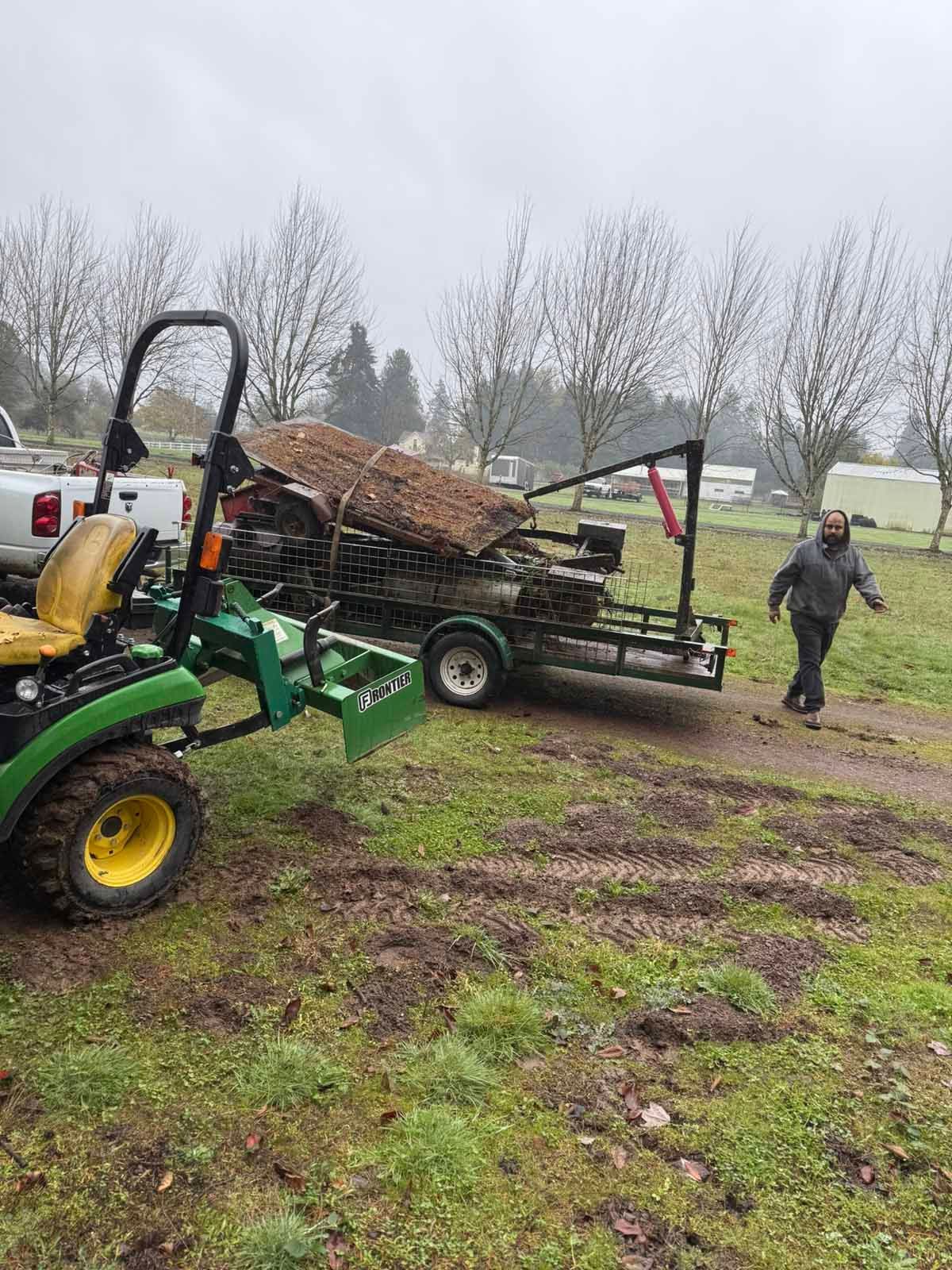 A green tractor and a trailer loaded with logs stand in a grassy field on an overcast day with a person walking nearby.