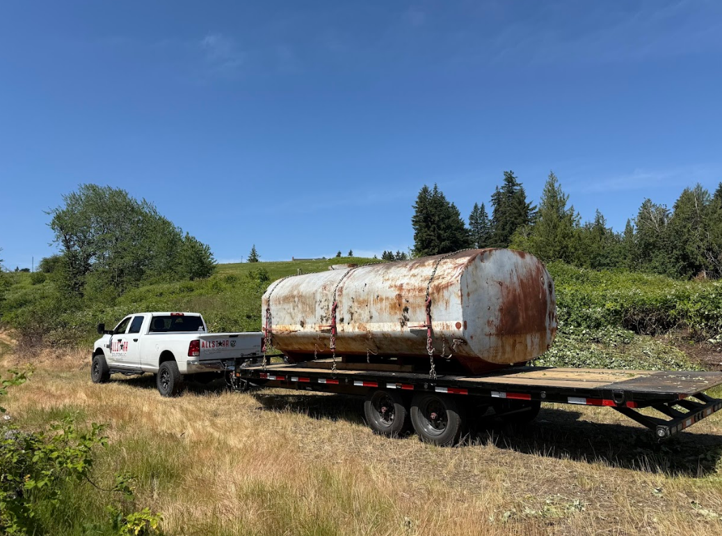 White pickup towing a rusty cylindrical tank on a flatbed trailer in a grassy field under a blue sky