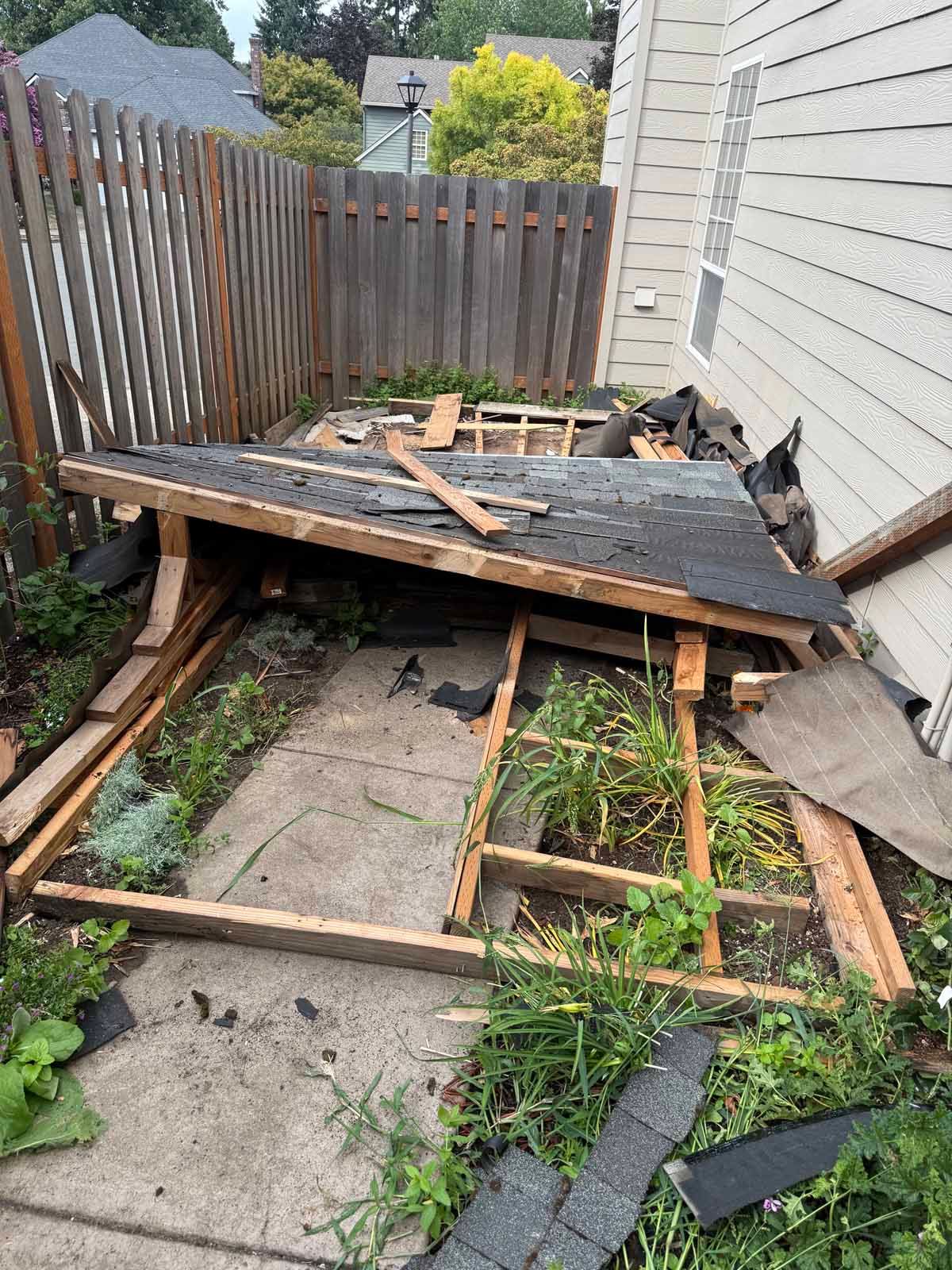 A collapsed wooden shed structure lying on its side on a concrete patio next to a house wall and wooden fence.