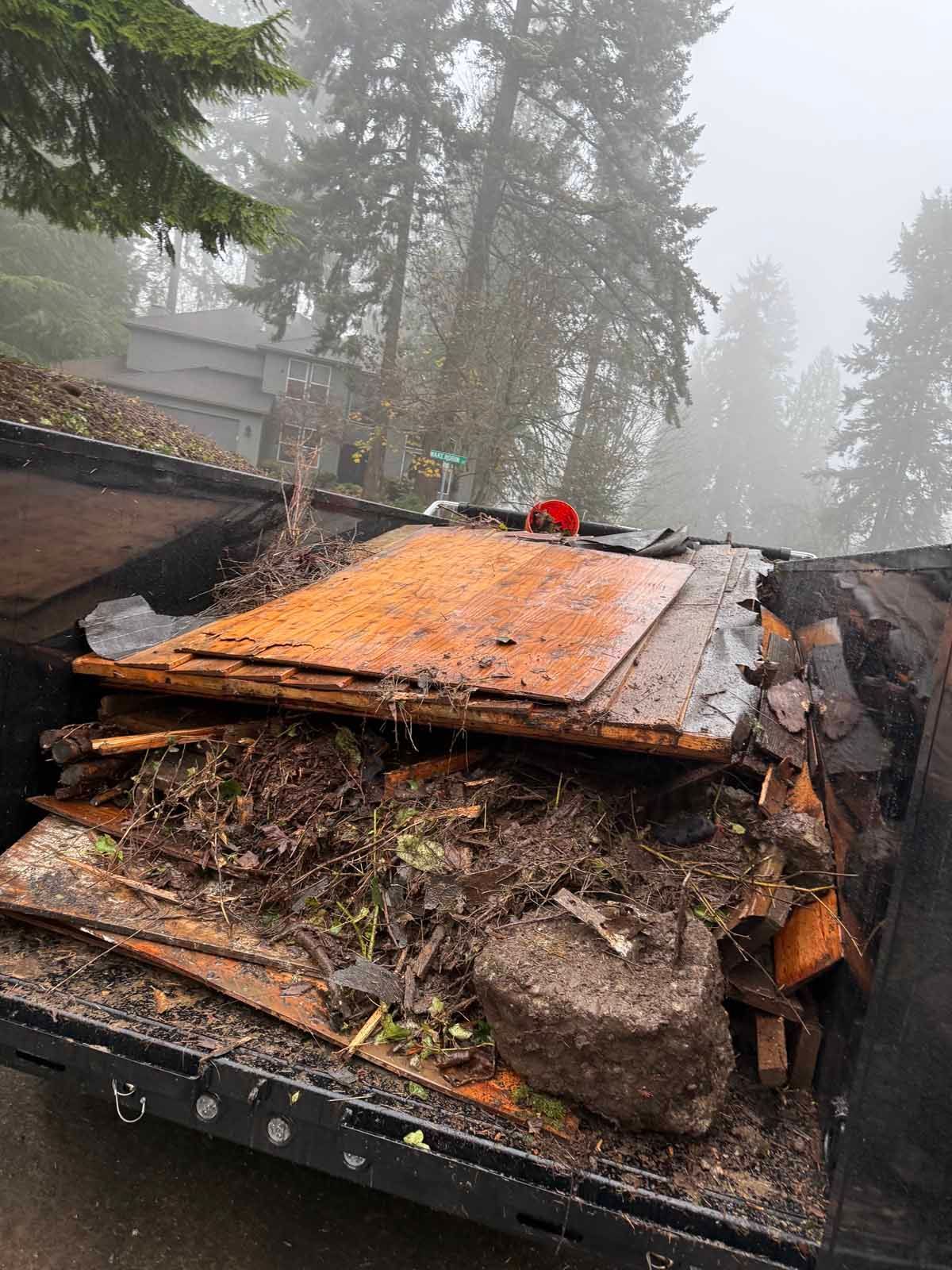 A trailer filled with discarded construction debris, including wood panels and dirt, parked on a foggy, wooded street.