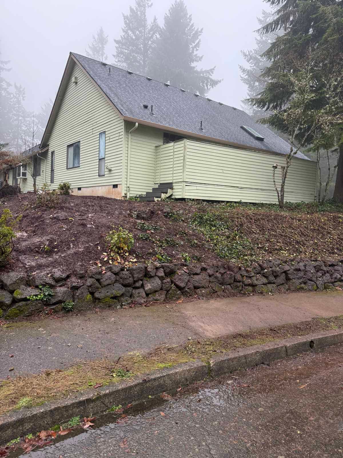 A pale green, single-story house with a grey roof sits on a sloped lot above a rock retaining wall and street.