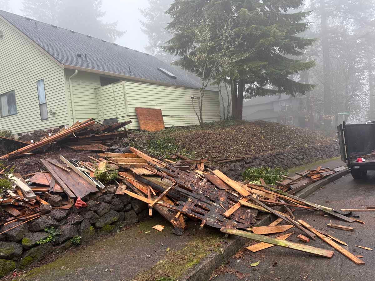 A pile of wooden debris and broken building materials sits on a stone wall next to a house under a foggy sky.