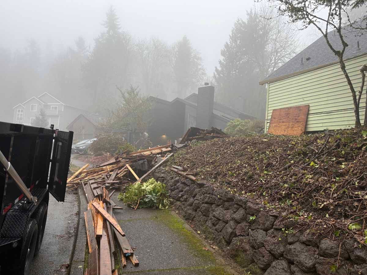 A pile of wooden debris lies on a driveway next to a stone retaining wall and a house on a foggy day.