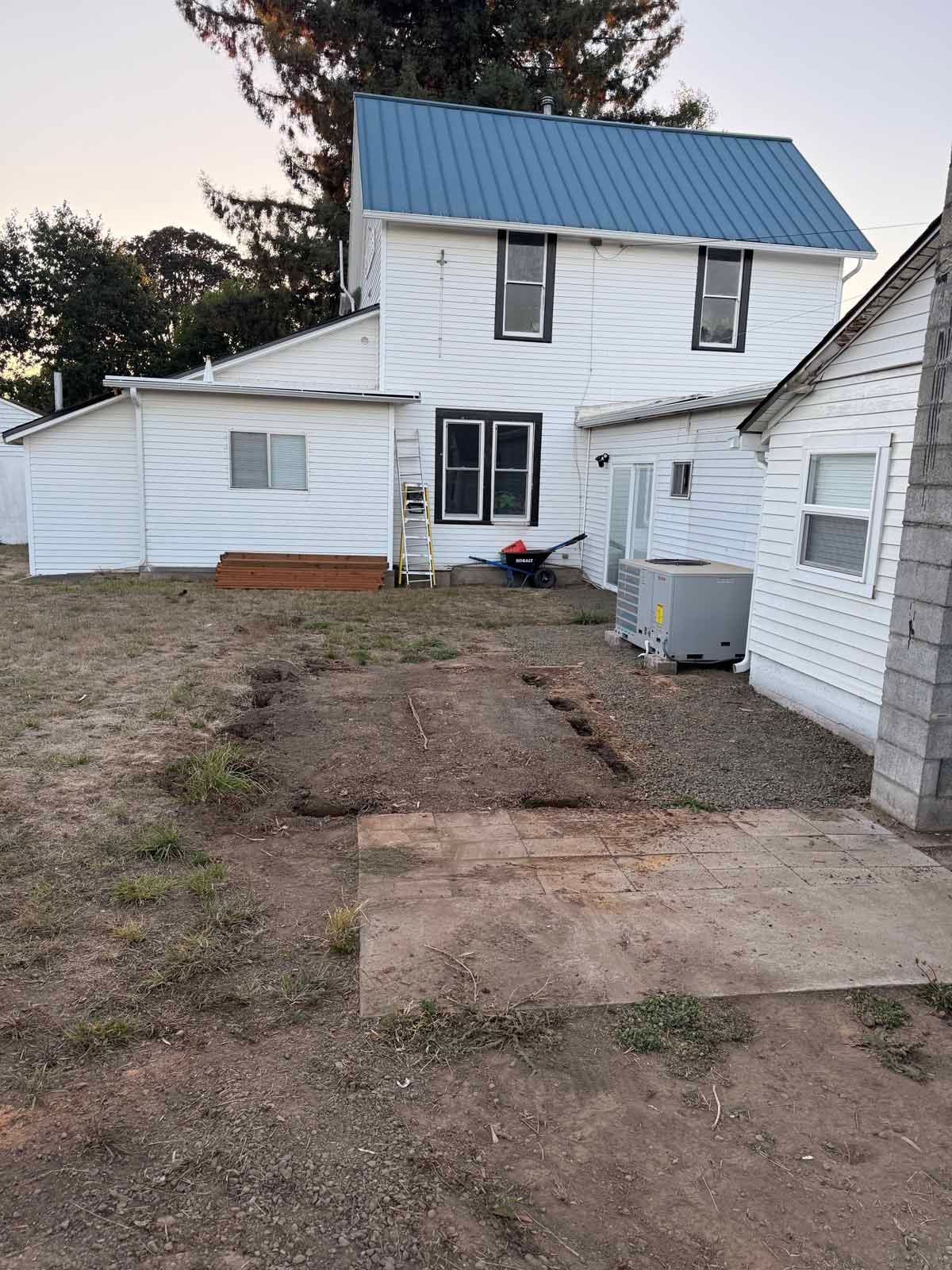 A white, two-story house with a blue metal roof, adjacent outbuildings, and a gravel yard with a concrete pad.