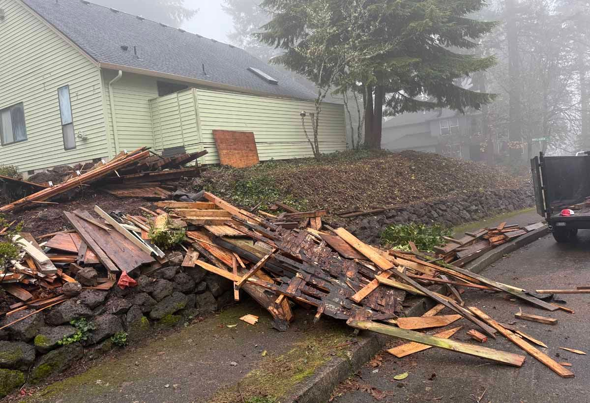 A house with light green siding behind a collapsed wooden fence and debris on a stone retaining wall in misty weather.