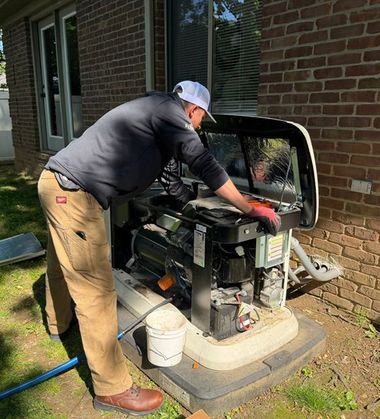 Technician kneeling beside a large HVAC unit, inspecting components with tools in a narrow outdoor space.