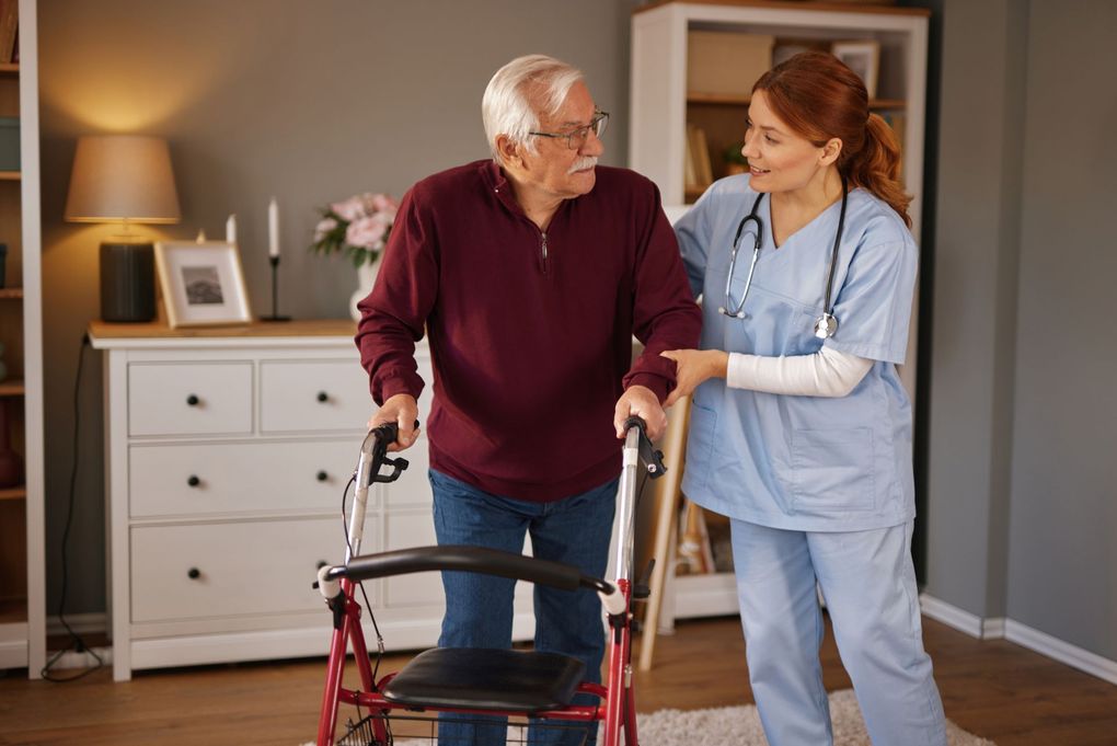 Caregiver helps an older adult walk with a walker in a home living room.