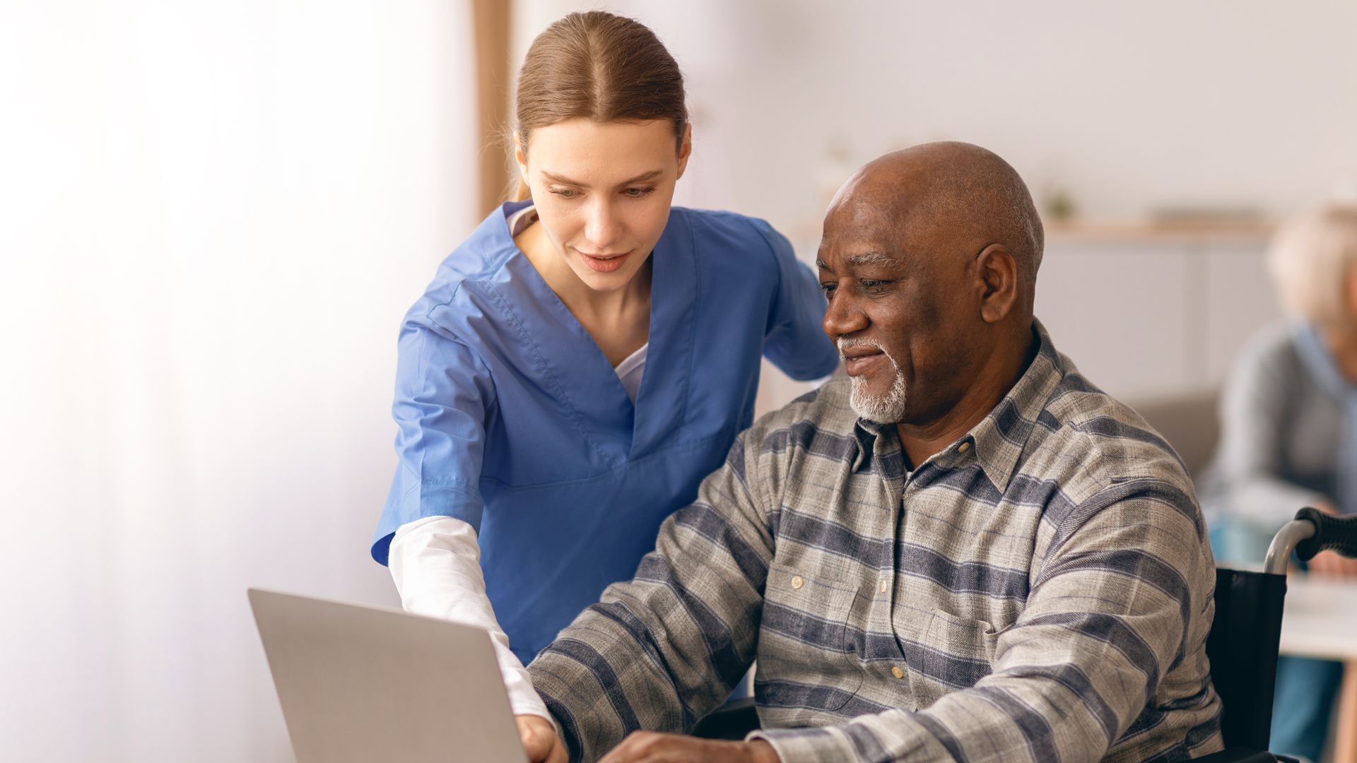 Caregiver helping a seated man use a laptop in a bright room Caregiver helping a seated man use a laptop in a bright room