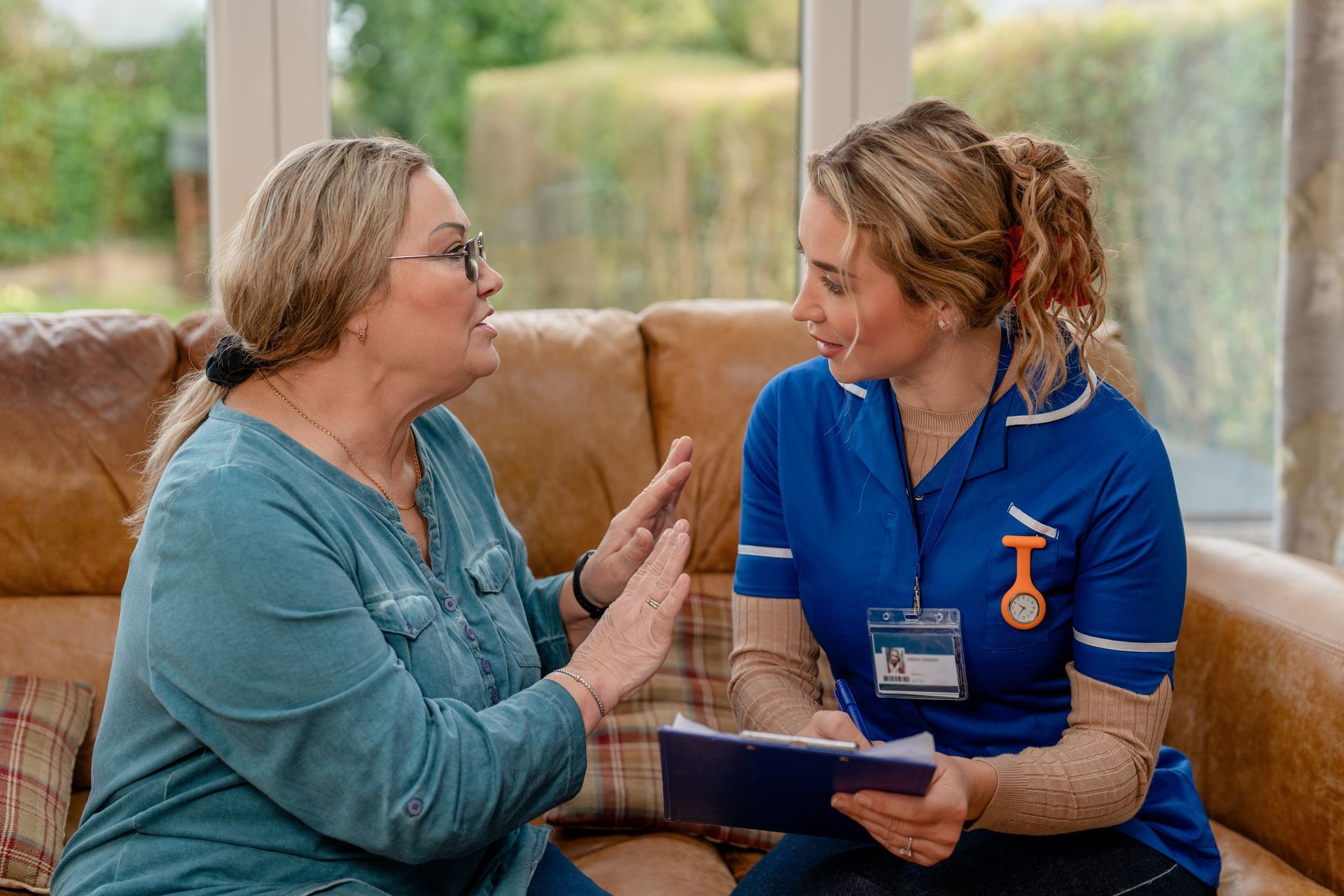 Two women talking on a couch, one in a blue uniform holding a tablet, in a bright living room Two women talking on a couch, one in a blue uniform holding a tablet, in a bright living room