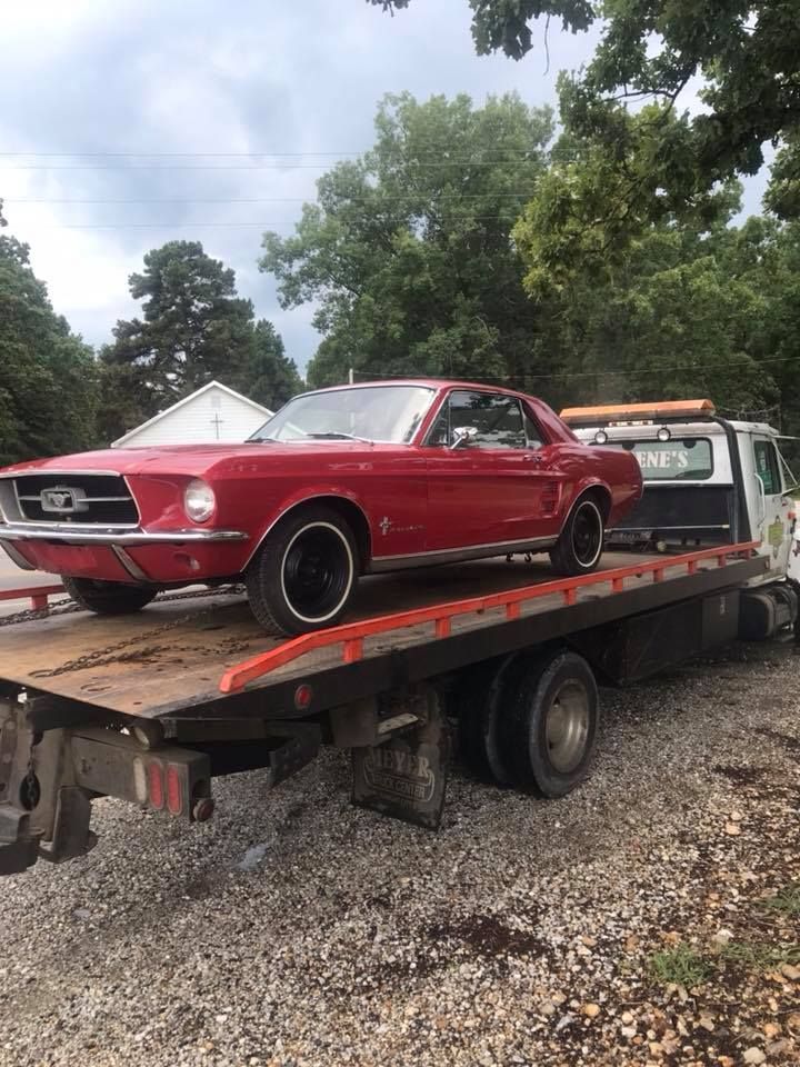 Red classic car strapped on a flatbed tow truck in a gravel lot with trees in the background