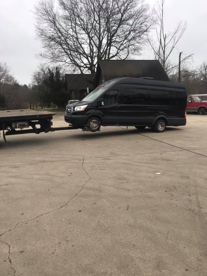 Black van hitched to a flatbed trailer on a concrete lot with a red vehicle and bare trees in the background