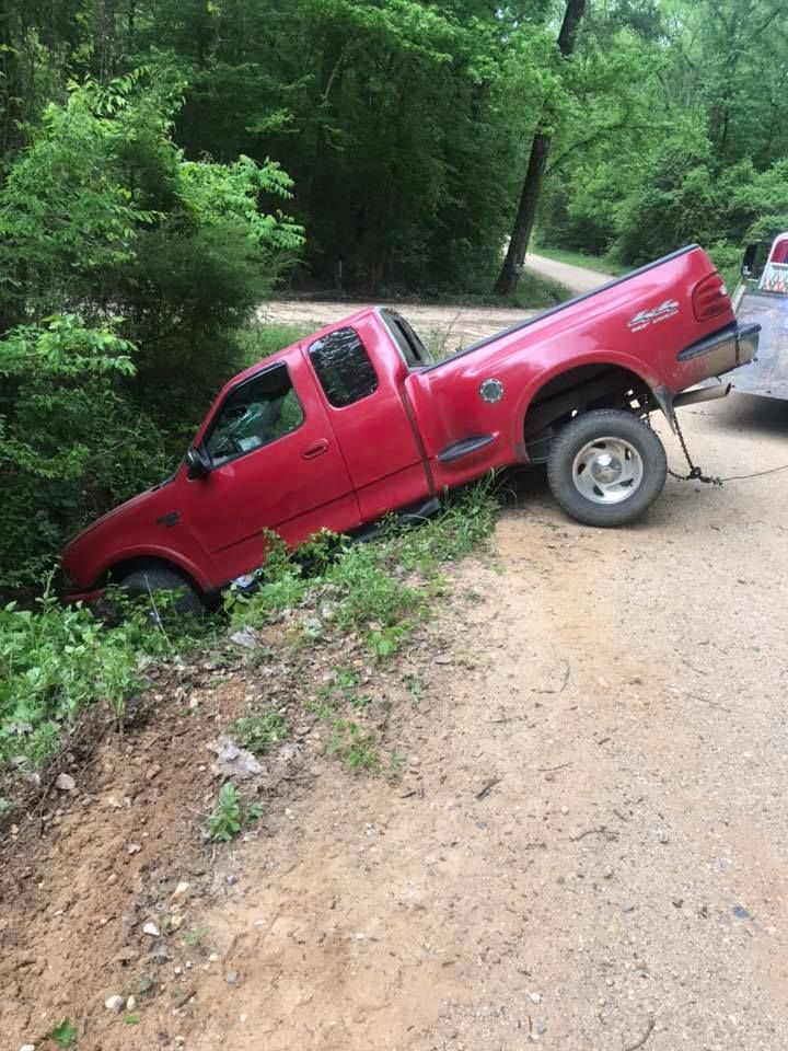 Red pickup truck stuck nose-down in a muddy roadside ditch beside a dirt road and trees