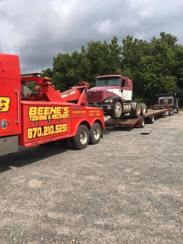 Red tow truck hauling an old pickup on a flatbed in a gravel lot under cloudy skies