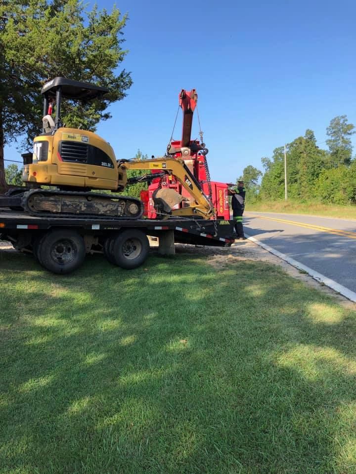 Mini excavator and red tow truck on a flatbed trailer beside a roadside grassy shoulder.