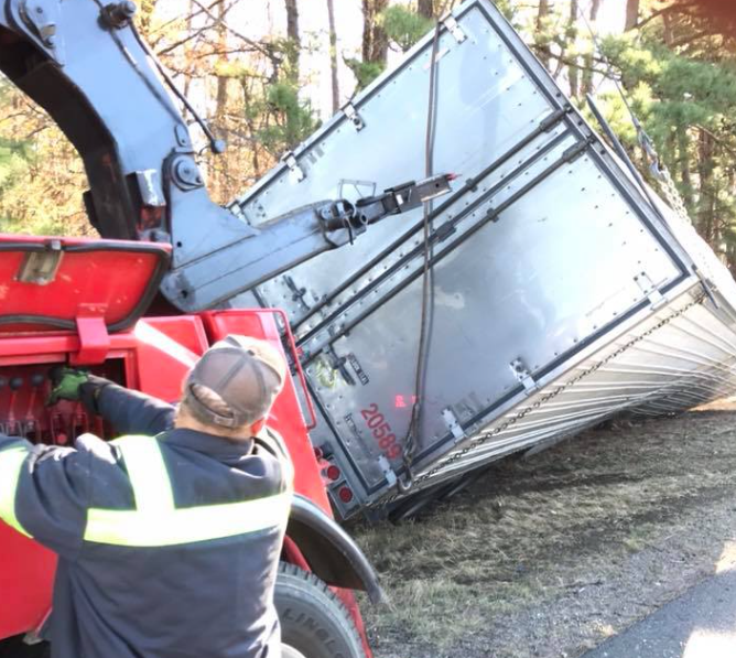 Tow truck driver beside an overturned silver trailer on a gravel roadside near trees