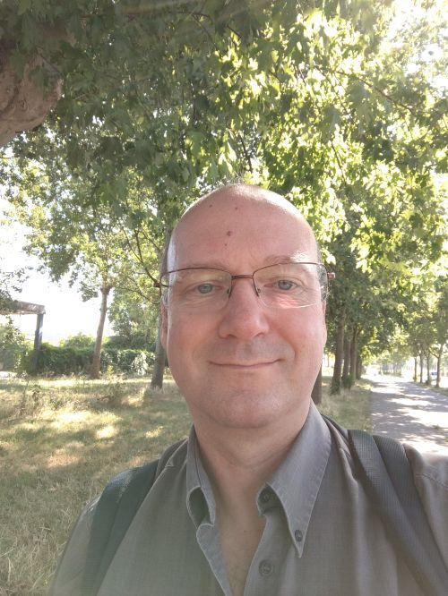 Een persoon met een bril en een lichtgekleurd overhemd met kraag staat lachend buiten onder lommerrijke bomen.