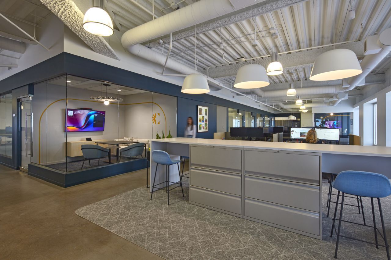 Modern office interior with open workspace, glass-walled meeting room, and overhead pendant lights.