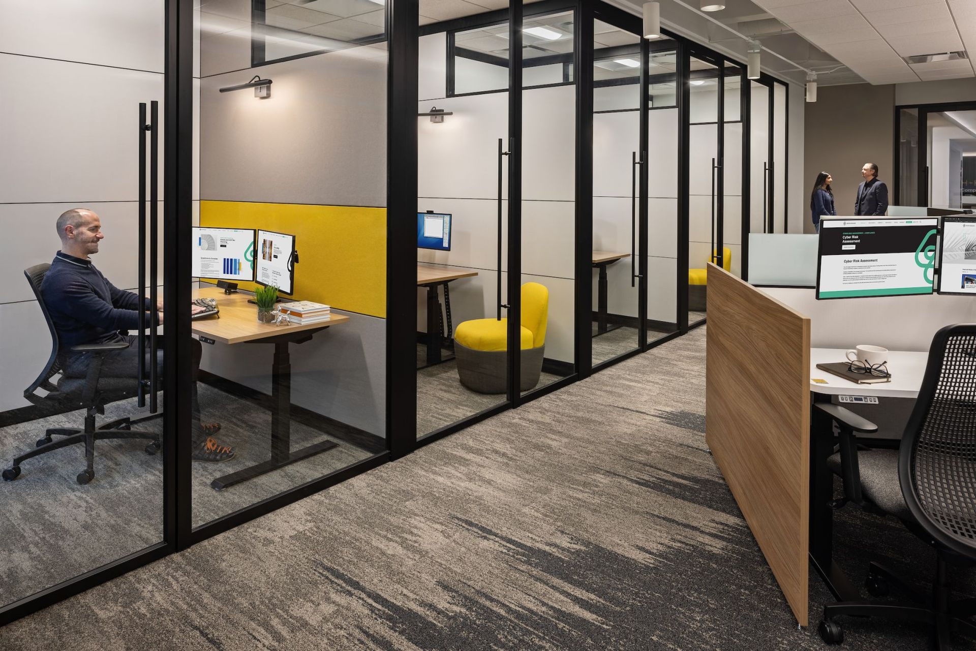 Modern office with glass-walled cubicles, desks, and a seated person working on a computer.