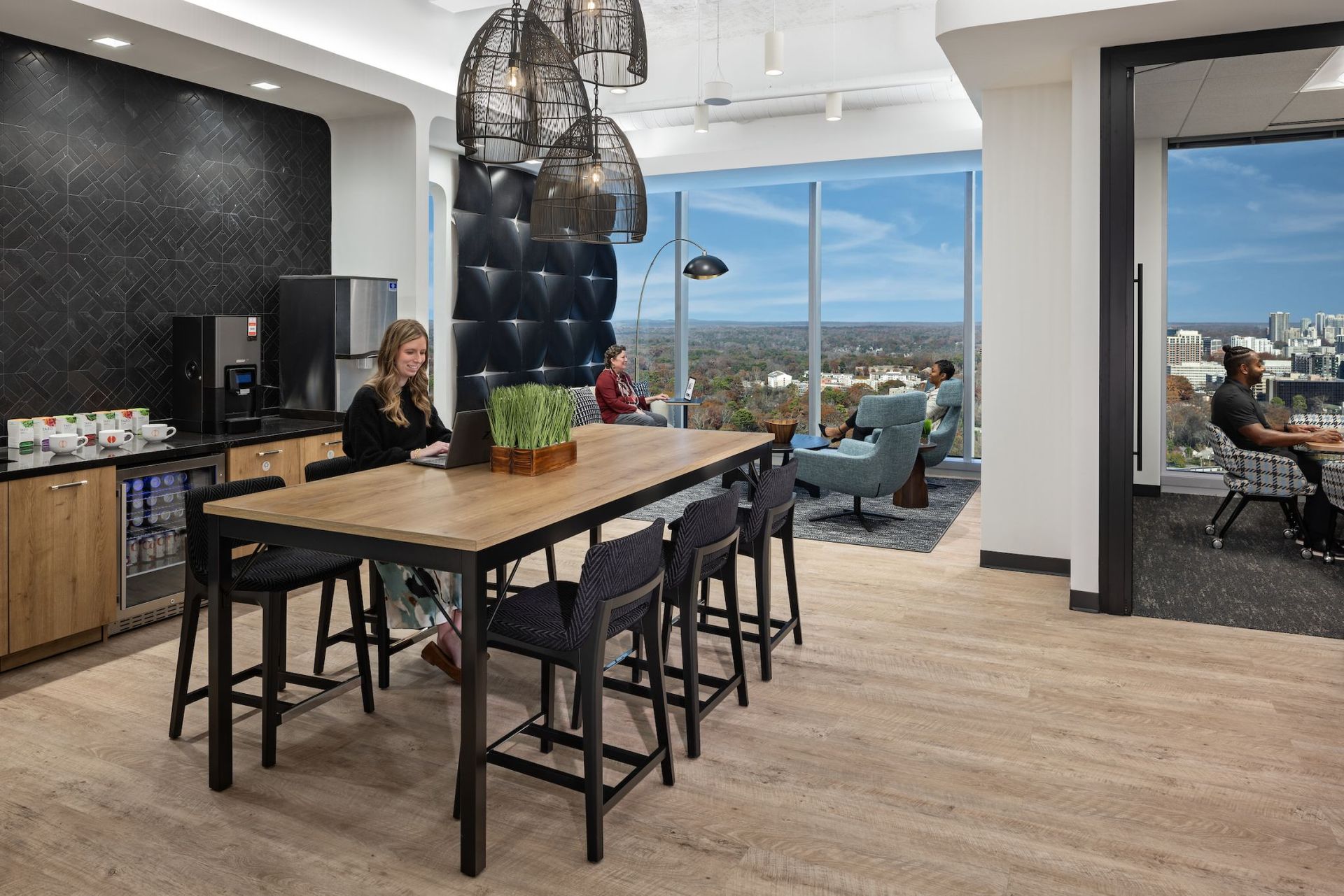 Modern office lounge with a long table, chairs, and coffee station. People are working and relaxing.