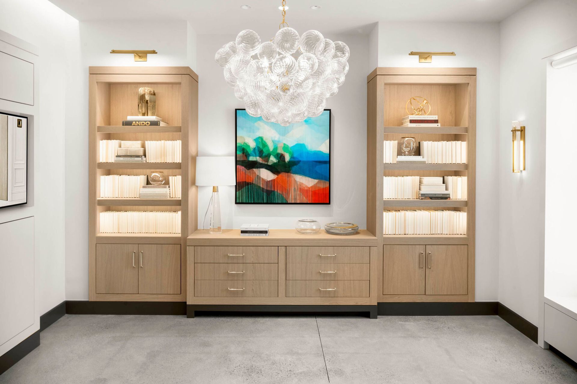 Home office with built-in cabinetry, artwork, and a chandelier. White walls, wood shelves, and a gray floor.