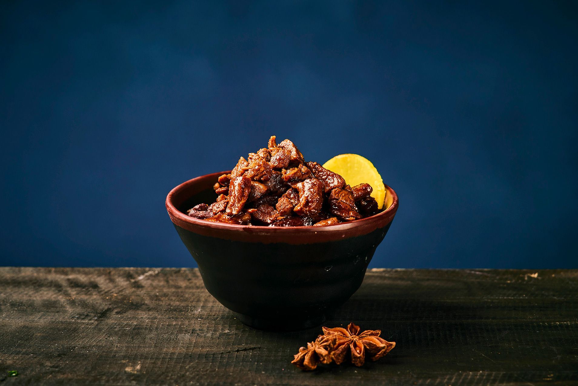 A bowl of saucy meat garnished with a lemon wedge on a dark table against a blue background