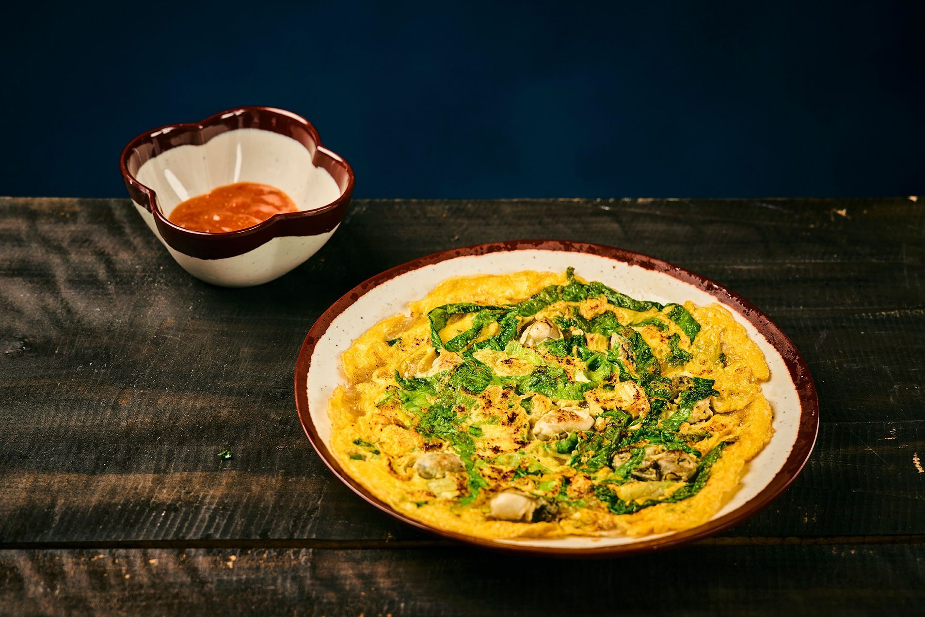 Yellow noodle dish on a plate with herbs, beside a small bowl on a dark wooden table