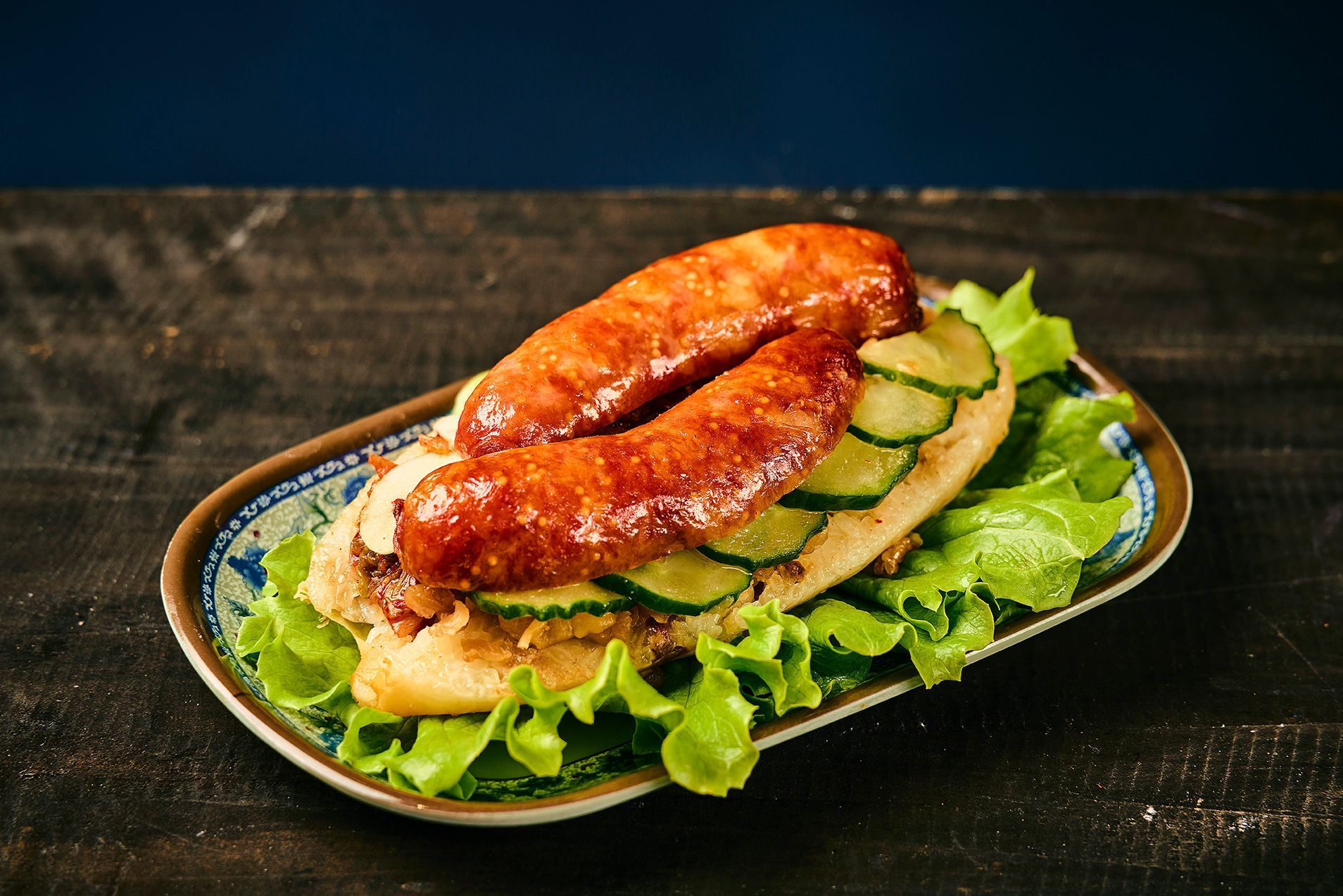 Roasted sausage sandwich on leafy greens with cucumber slices on a dark plate