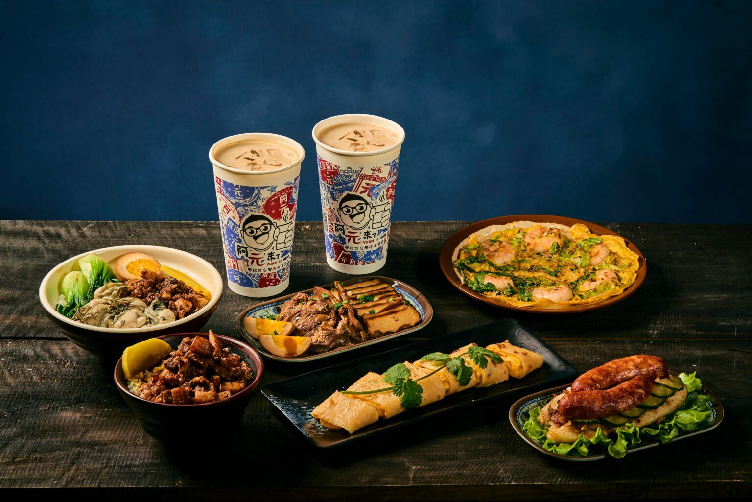 Assorted takeout dishes and two iced drinks on a dark table against a blue backdrop