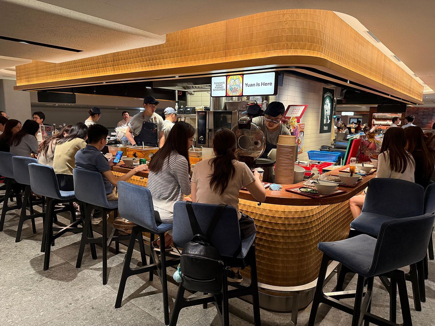 Crowded sushi bar with people seated at a curved counter under warm wood lighting