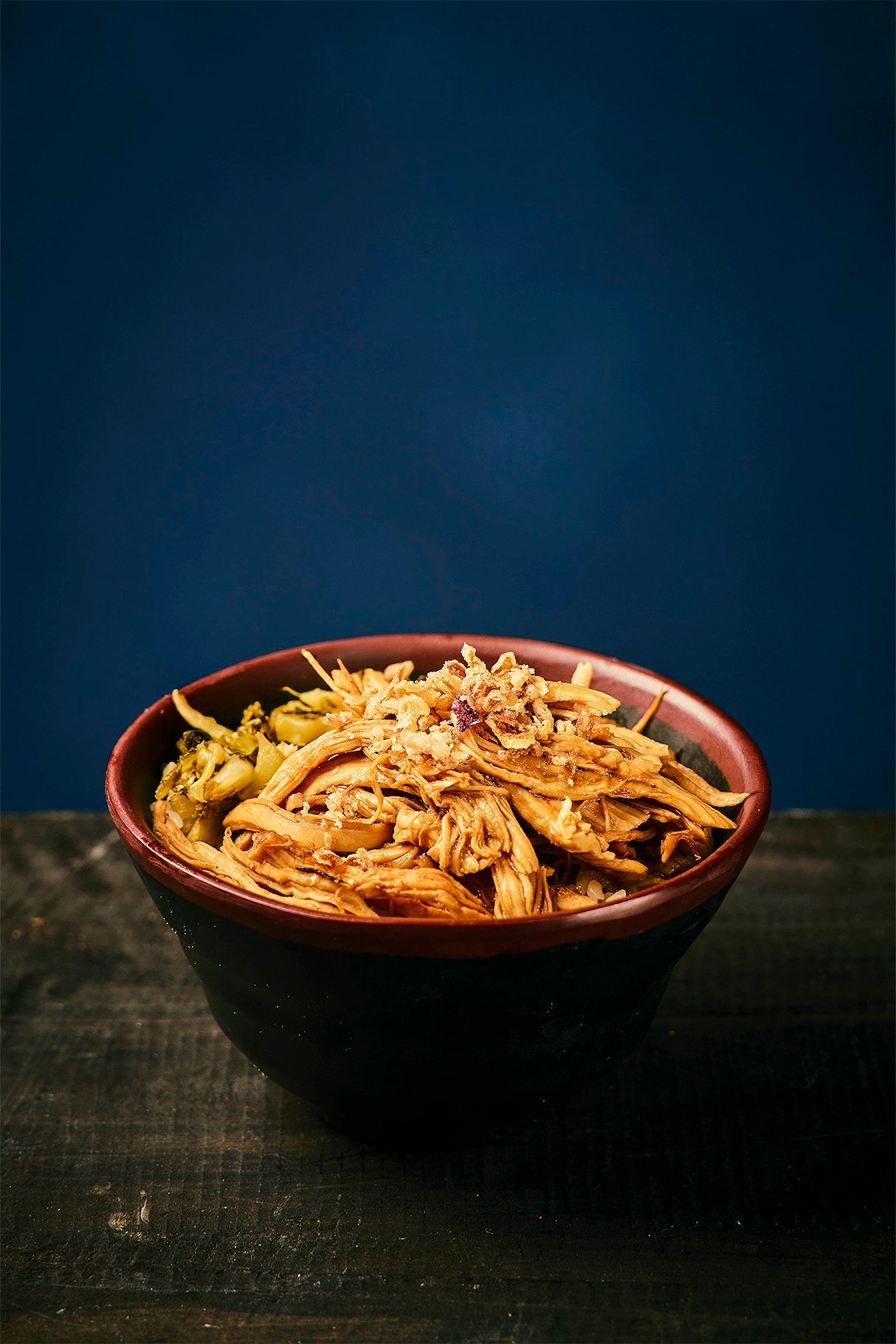 Bowl of shredded food topped with seasoning on a dark table against a blue background