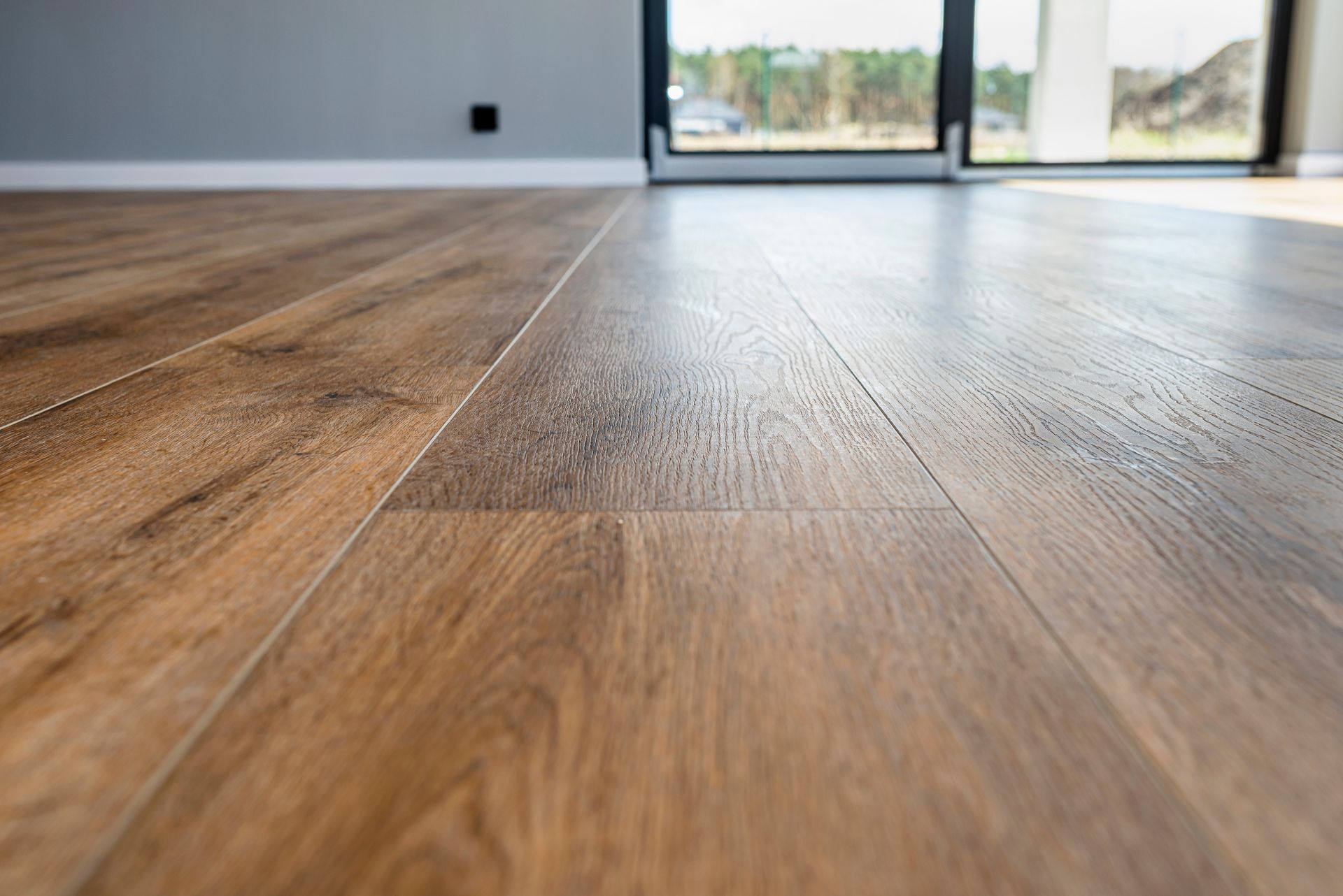 Close-up of glossy, light brown wood flooring in a room with a large window and gray wall.