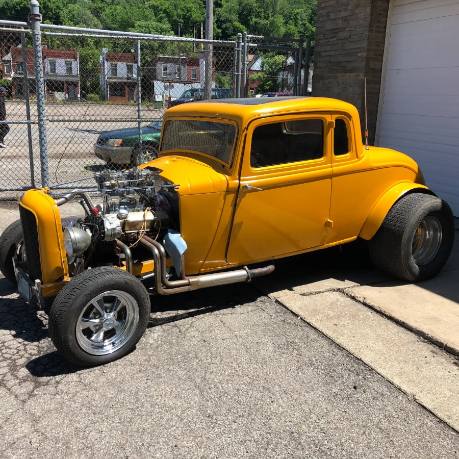 Yellow hot rod coupe parked in a driveway, with exposed engine and black rear wheels.