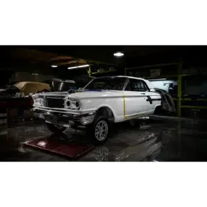 White classic car in a dim garage, parked on a lift with front-end repairs in progress