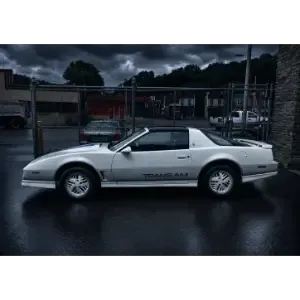 Silver sports coupe parked on a wet street, side view, with dark cloudy sky and fenced lot behind it
