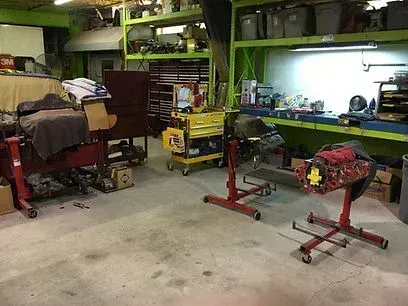 A cluttered garage workshop with tools, a workbench, and two engine stands on a concrete floor