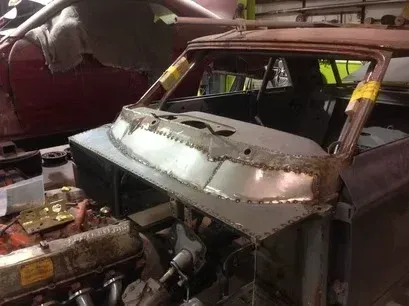 Rusty car body under repair in a workshop, with stripped front and exposed engine.
