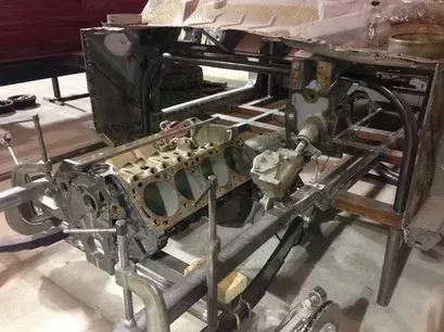 Disassembled car engine and chassis frame in a workshop, with exposed metal parts and tools.